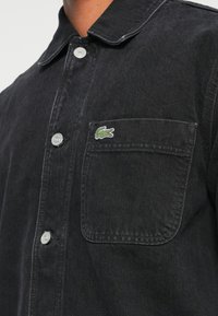 Camicia in denim nero con bottone frontale, colletto e taschino sul petto. Presenta un piccolo logo verde Lacoste sulla tasca.