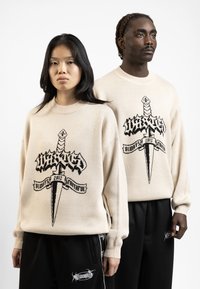 Beige gebreide trui met zwart grafisch ontwerp van een zwaard en tekst. Model in zwarte broek met witte accenten staat naast een ander model.