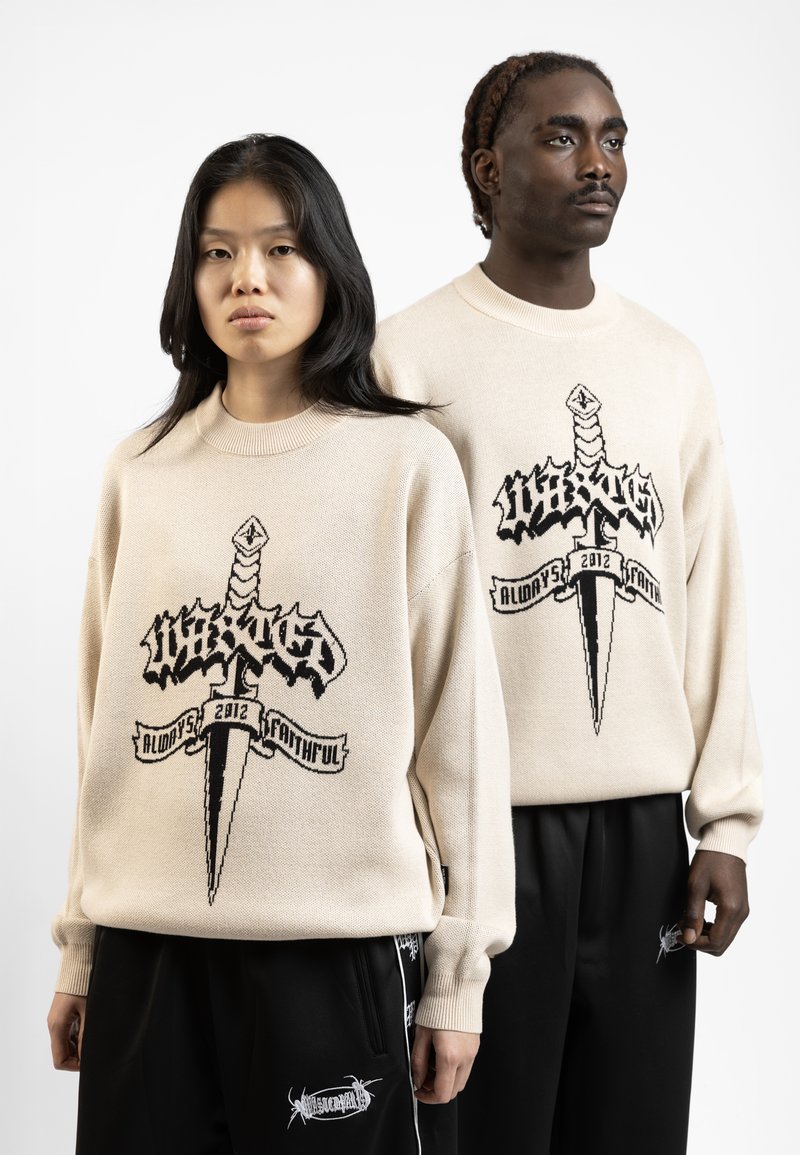 Beige gebreide trui met zwart grafisch ontwerp van een zwaard en tekst. Model in zwarte broek met witte accenten staat naast een ander model.