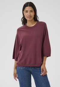 Maglione a maglia color marsala con scollo rotondo, maniche a tre quarti e una vestibilità ampia. Abbinato a jeans in denim blu. Texture liscia.