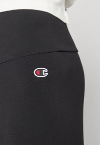 Fekete sportleggings sima felülettel és testhezálló szabással, az oldalon egy kicsi piros-fehér hímzett logóval.