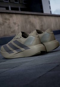 Ζευγάρι μπεζ αθλητικά παπούτσια Adidas με σκούρες ρίγες και παχιά μαξιλαρωμένη σόλα τοποθετημένα σε υπαίθριο πεζοδρόμιο.
