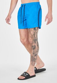Pantaloni da nuoto blu con cordino nero e strisce laterali, abbinati a infradito nere con logo, mostrati su una gamba coperta di tatuaggi.