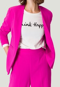 Blazer fucsia sobre una camiseta blanca con el texto "Think Happy", combinado con pantalones fucsias a juego, mostrando un ajuste entallado y elegante.