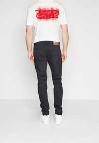 Valkoinen t-paita, jossa on punainen graafinen kuvio selässä, yhdistettynä istuviin mustiin farkkuihin ja valkoisiin lenkkitossuihin. Yksinkertainen, moderni tyyli.