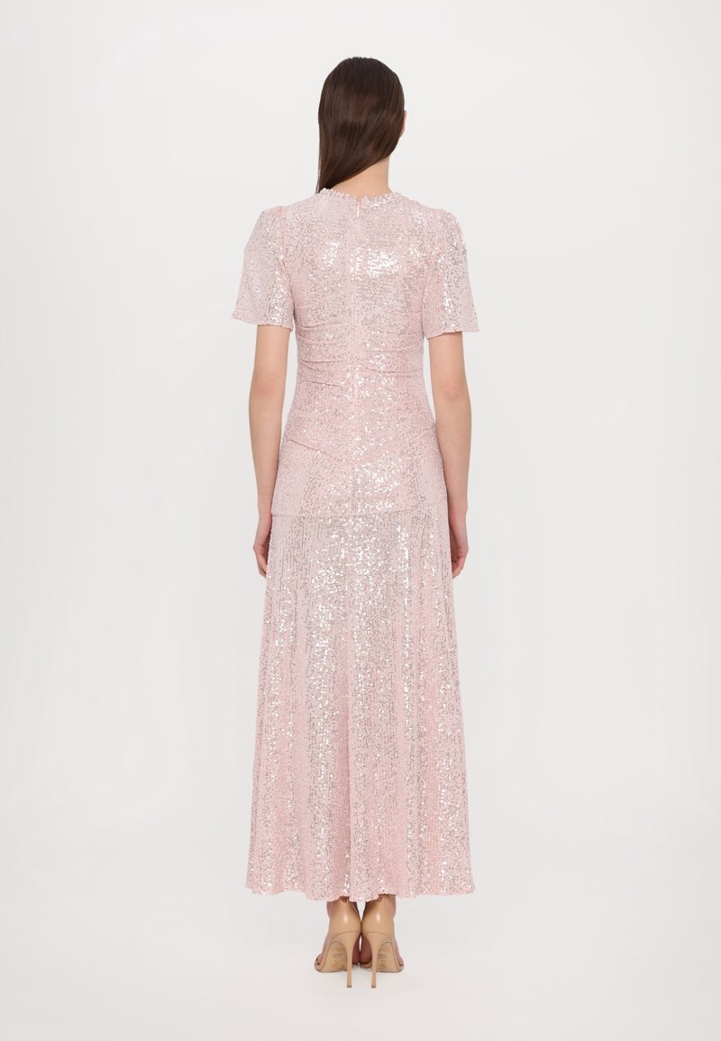 Femme aux longs cheveux foncés, vêtue d'une robe longue rose clair à manches courtes et paillettes, et de talons hauts beiges, debout, tournée de dos.