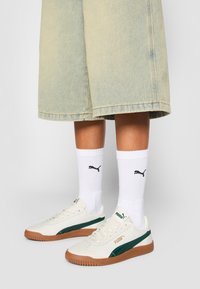 Vita sportskor med gröna och guldiga detaljer, med en texturerad, brun gummisula. Outfit inkluderar vita strumpor och ljusa denimshorts.