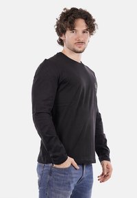 T-shirt nera a maniche lunghe in cotone, con scollatura rotonda e un piccolo logo sul petto. Indossata con jeans blu.