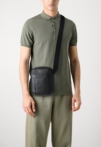 Borsa a tracolla sintetica nera con pannello frontale testurizzato e logo. Caratterizzata da una tracolla regolabile e una finitura liscia. Forma rettangolare compatta.