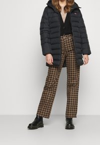 Kvinna klädd i svart puffjacka, svart knäppt kavaj, bruna och svarta houndstooth-byxor och svarta ankelskor som står framför en vit bakgrund.