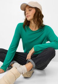 Femme portant une casquette beige, une chemise à manches longues verte, un jean noir retroussé, des chaussettes beiges et des bottes couleur tan, assise en tailleur sur un sol blanc.