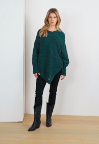 Poncho lavorato a maglia color teal con un motivo diagonale, indossato sopra pantaloni neri aderenti e stivali da cow-boy neri con tacco affusolato.