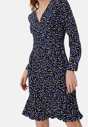 Mujer con un vestido cruzado negro con estampado floral en blanco y azul y mangas largas, de pie sobre un fondo liso.