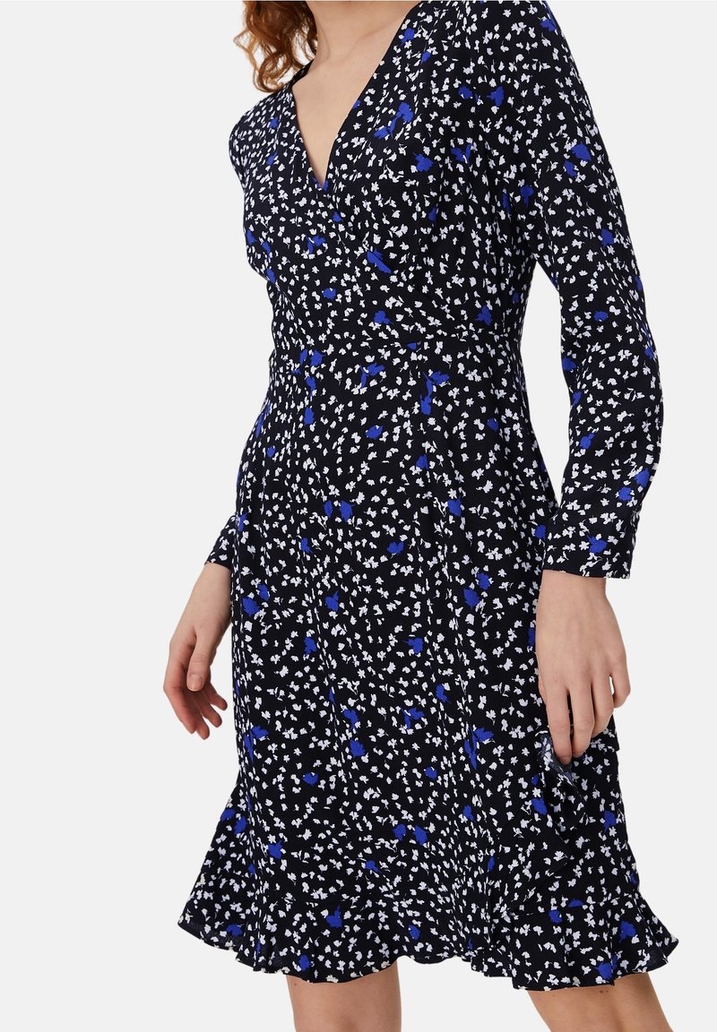 Femme portant une robe portefeuille noire à imprimé floral blanc et bleu avec manches longues, debout devant un fond uni.
