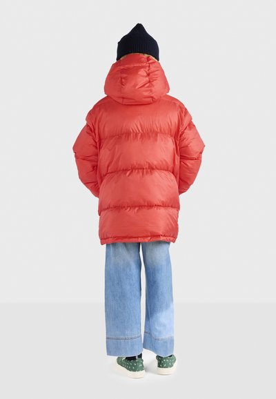 Person in einer hellroten Puffjacke mit Kapuze, blauen, weit geschnittenen Jeans, grünen, gemusterten Turnschuhen und einer schwarzen Mütze, der mit dem Rücken zur Kamera steht.