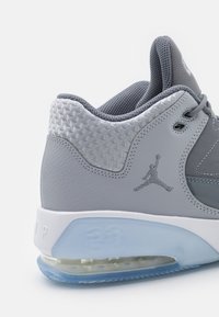 Zapatilla deportiva gris con parte superior de cuero texturizado, detalles de malla, amortiguación de aire visible y suela blanca. Presenta el logo Jumpman en gris.