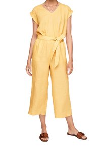 Femme portant une combinaison courte jaune à manches courtes avec une ceinture nouée à la taille et des sandales marron ouvertes, debout sur un fond blanc.