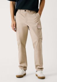 Beige cargobroek gemaakt van stevig stof, met meerdere zakken, een knoopsluiting en een getailleerd ontwerp. Draagt bij witte sneakers.