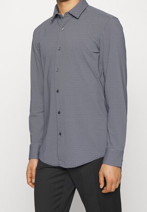 Mężczyzna ubrany w koszulę na guziki z długim rękawem o małym czarno-białym geometrycznym wzorze oraz czarne spodnie na jednolitym tle.