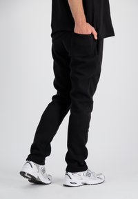 Sorte joggerbukser har et tætsiddende design med en glat tekstur, accentueret af sidelommer. Båret sammen med hvide New Balance sneakers.