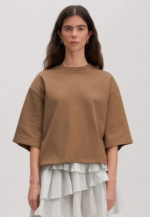 Frau mit langen dunklen Haaren trägt ein übergroßes braunes Kurzarmoberteil über einem mehrlagigen, gerafften, gestreiften weißen Rock vor einfachem Hintergrund.