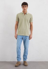 Lichtgroene poloshirt met een drieknoopsluiting, gecombineerd met lichtblauwe jeans en bruine suède laarzen. Slim fit, zachte textuur, minimale branding.