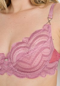 Soutien-gorge en dentelle rose pâle avec des bords festonnés et des motifs géométriques. Bretelles fines réglables avec détail en métal sur l'épaule.