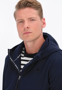 Tamnoplava zip-up dukserica s teksturiranim unutrašnjim dijelom, s prugastom crno-bijelom majicom ispod i logotipom na prsima.