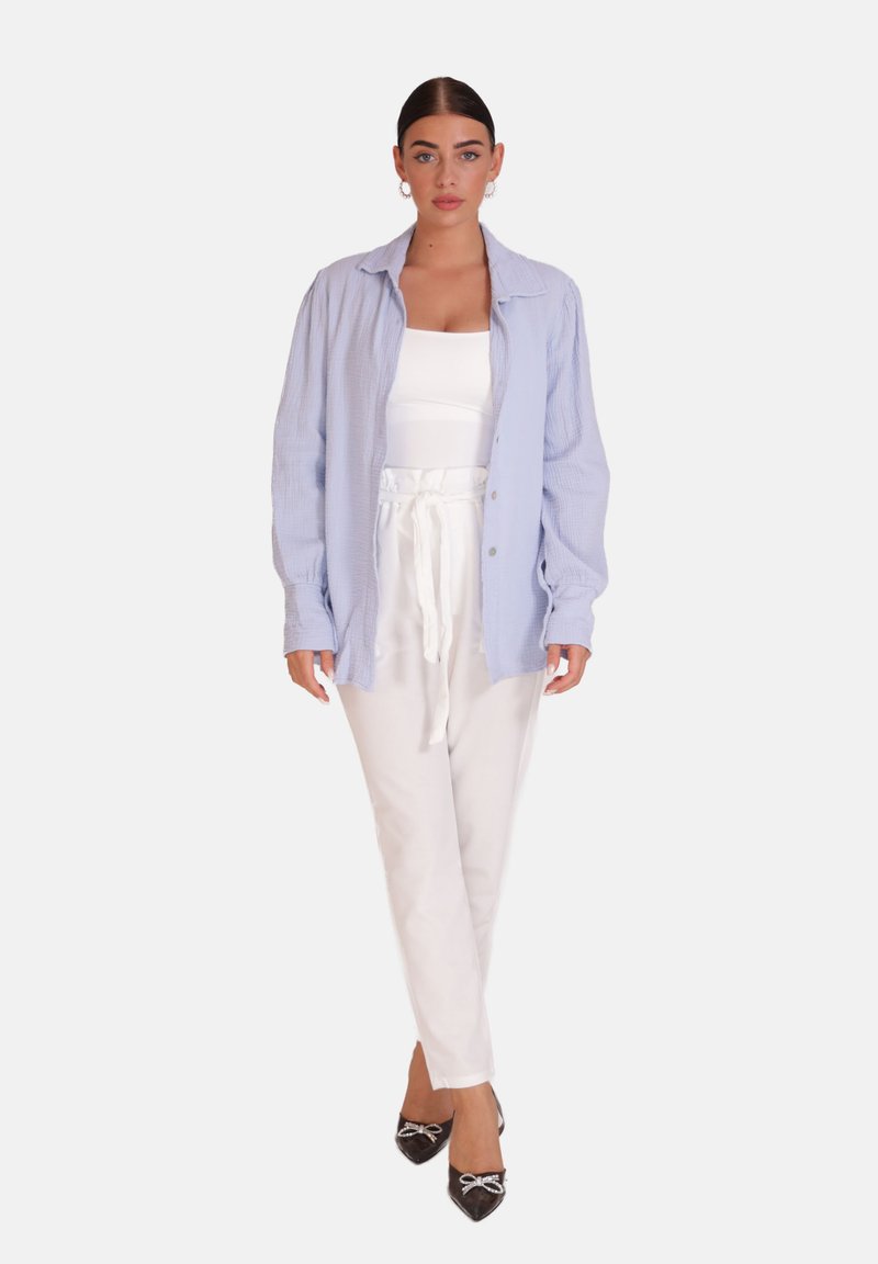 Vrouw draagt een lichtblauw overhemd over een wit tanktop, witte broek met hoge taille en trekkoord, en zwarte puntige schoenen met strikdetail.