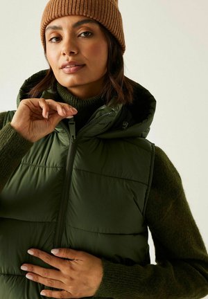 Kobieta w beżowej czapce i zielonej puchowej kamizelce na ciemnozielonym swetrze, zapinająca kamizelkę na suwak na neutralnym tle.