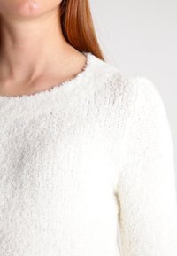 Gros plan sur une personne portant un pull doux et texturé blanc avec un col rond, sur un fond clair uni.