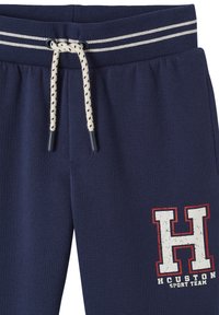 Marineblaue Sweatpants mit einem weißen gestreiften Bund. Sie sind mit einem elastischen Kordelzug und dekorativem Zubehör ausgestattet. Beinhaltet ein aufgedrucktes "H"-Logo.