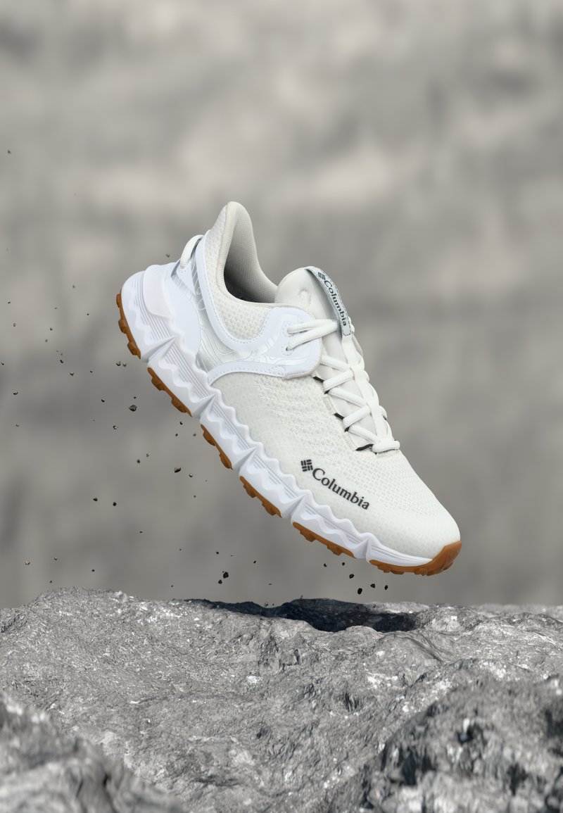 Fehér Columbia sportcipő texturált talppal, köves talaj felett lebegve, apró homokszemcsékkel a levegőben.