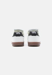 Λευκά αθλητικά παπούτσια με καστανές λαστιχένιες σόλες, μαύρες λεπτομέρειες στη φτέρνα και το «1973» ανάγλυφο στο πίσω μέρος. Ομαλό υλικό με απαλό εσωτερικό επένδυση.