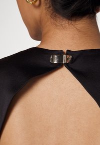 Gros plan sur la nuque et le haut du dos montrant un vêtement noir attaché par une boucle dorée brillante et une boucle d'oreille en anneau doré à l'oreille gauche.