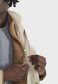 Beige Außenjacke mit glatter Textur, ausgestattet mit einem Frontreißverschluss und Knopfverschlüssen. Braune Fleece-Innenschicht sorgt für Wärme. Graues Hemd darunter.