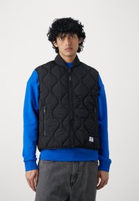 Gepolsterte schwarze Weste mit Reißverschluss, Seitentaschen und hohem Kragen, die über einem blauen Sweatshirt getragen wird. Graue Jeans runden das Outfit ab.