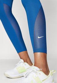 Siniset urheilulegginsit, joissa on verkkosivupaneelit ja valkoinen Nike-logo. Yhdistetty valkoisiin kenkiin, joissa on keltaisia korostuksia.