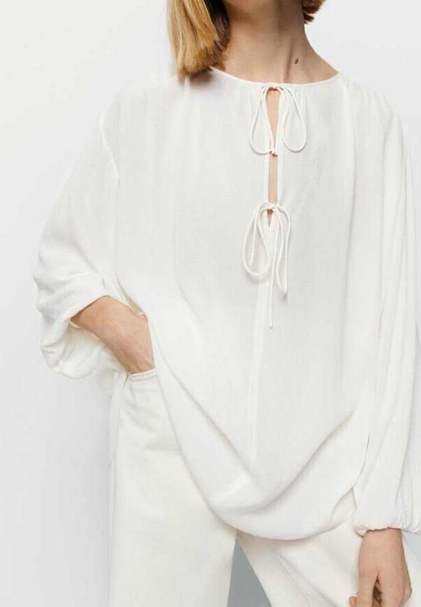 Femme portant un chemisier blanc ample avec deux liens au niveau du col et de longues manches ballon, assorti à un pantalon blanc.