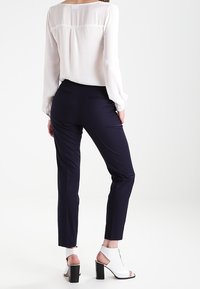 Vit långärmad blus med en subtil söm bak, matchad med marinblå slim-fit byxor och vita öppna blockklacksskor.