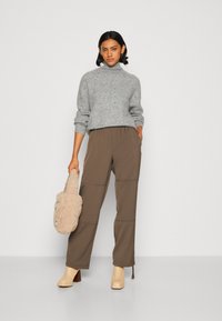 Pull gris clair à col montant, associé à un pantalon élastiqué marron et des bottes à talons beiges. Tenant un sac à main en fourrure beige.