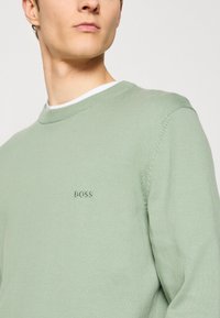 Šviesiai žalias megztinis su raukšlėta apykakle, pasižymintis mažuoju siuvinėtu "BOSS" logotipu ant krūtinės ir lygiu paviršiumi.