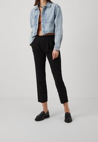 Veste en denim bleu clair avec deux poches poitrine, portée sur un crop top marron texturé, associée à un pantalon noir fuselé et des mocassins noirs brillants.