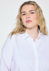 Bílá košile na knoflíky s límečkem, vyrobená z hladké látky; má jednoduchý design a mírný lesk. Model má vlnité blond vlasy.