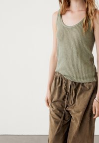 Femme portant un haut en tricot sans manches vert olive et un pantalon ample marron avec cordon, debout devant un fond clair uni.