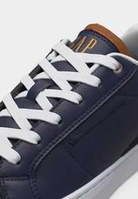 Baskets en cuir bleu marine avec lacets blancs, doublure marron sur la languette et logo "GAP" doré sur la languette.