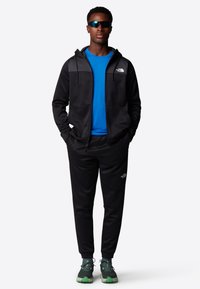 Black zip-up hoodie with gray accents, blue t-shirt, black sweatpants, and green-trimmed running shoes. Reflective sunglasses complete the look.