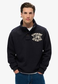 Granatowy sweter z wysokim kołnierzem, zapięcie na guziki i kremowy napis "SUPERDRY JPN ATHLETIC" na klatce piersiowej. Materiał z mieszanki bawełny.