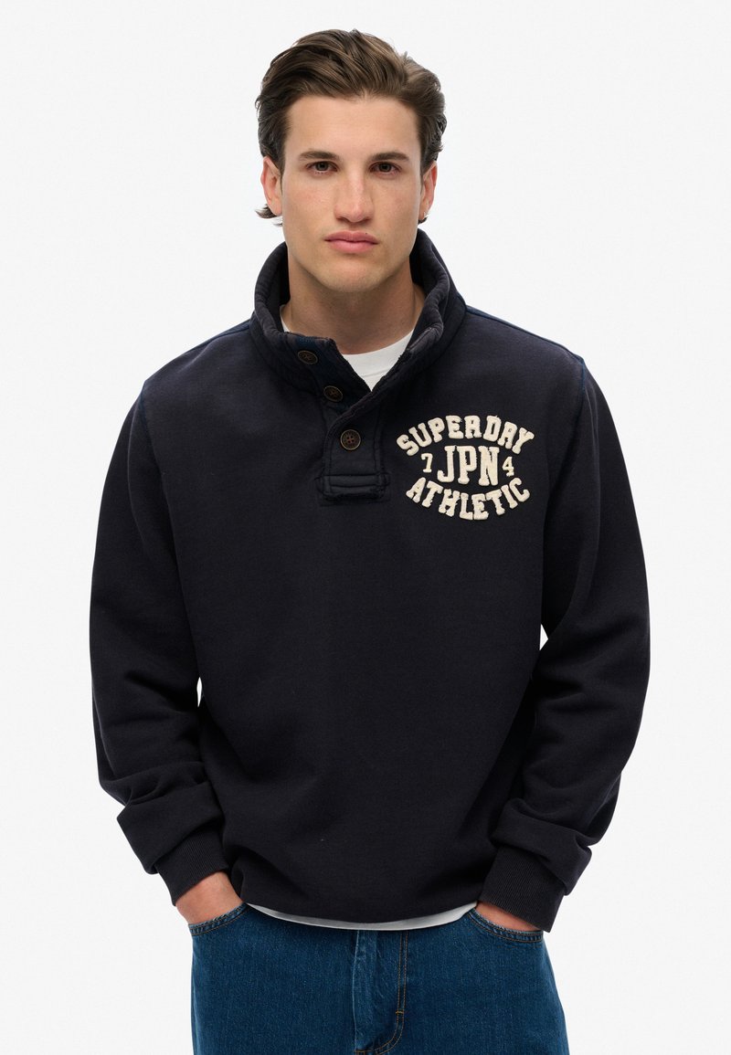 Granatowy sweter z wysokim kołnierzem, zapięcie na guziki i kremowy napis "SUPERDRY JPN ATHLETIC" na klatce piersiowej. Materiał z mieszanki bawełny.