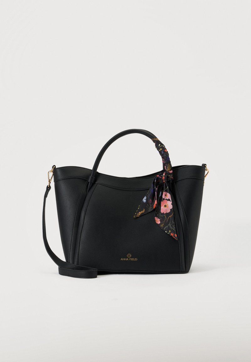 Sac à main en cuir noir avec double poignées, un foulard fleuri noué sur une poignée et une bandoulière amovible sur fond uni.
